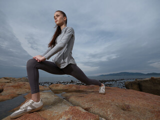 Young female athlete stretching on rocks by the sea, dressed in sportswear, showcasing...