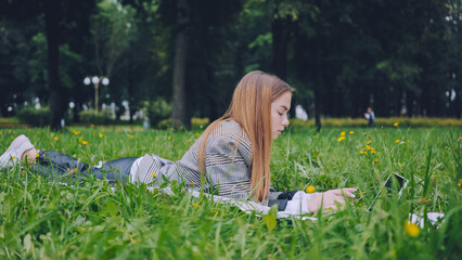 College student typing on laptop while relaxing on lush grass, embracing outdoor workspace with natural surroundings