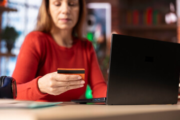 Confident woman holds her card while she pays for the online order, using laptop at home office to...