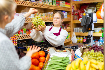 Young friendly salesgirl working at counter in grocery, taking grape cluster from female customer for weighing..