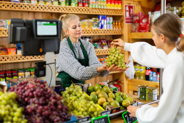 Friendly saleswoman working behind counter in grocery, taking grape cluster from female customer for weighing..