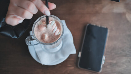 Smiling woman stirring marshmallow topped hot chocolate, relaxing at cafe with smartphone resting nearby