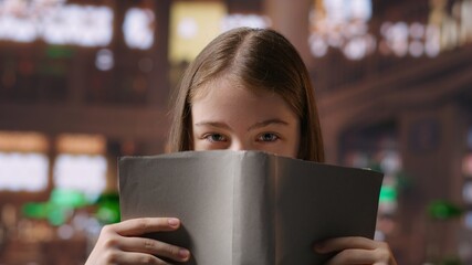 Funny preteen student covering her face with a book in the school library, playing around and...