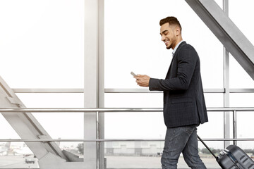Fototapeta premium Handsome arab guy using smartphone while walking with suitcase at airport terminal, young middle eastern man browsing mobile internet on cellphone while going to flight boarding, side view