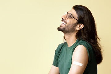 Fototapeta premium Joyful middle-eastern young guy with adhesive medical band on shoulder resting on chair after immunization, beige studio background, panorama with copy space. Vaccination while COVID-19 pandemic