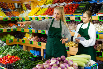 Young trainee salesgirl at greengrocery store listening attentively to experienced female colleague and making notes. First days of work concept