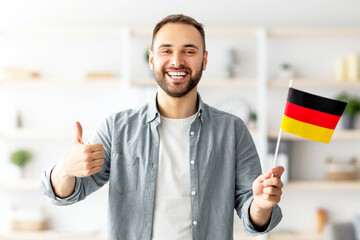 Happy Caucasian man showing thumb up and flag of Germany, posing and smiling at camera indoors....