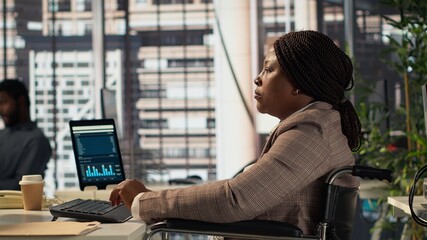 Black manager wheelchair user reviewing the financial data for the monthly reports, ensuring an accurate revenue balance. Woman with disability solving business operations. Camera A.