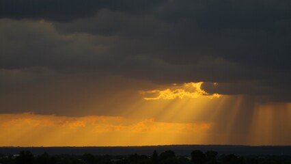 Fototapeta premium A dramatic sky at dusk with golden sunbeams breaking through thick gray clouds, casting radiant streaks of light over an amber-hued horizon.