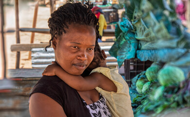 African woman vendor holding baby selling vegetables from a wooden shack stall in the city, on the...