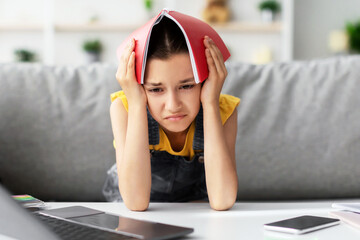 School Stress Concept. Tired upset student girl holding copybook on head sitting on sofa at desk,...
