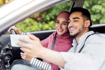 Cheerful glad millennial arab man and woman in hijab enjoy journey in new car and take selfie on smartphone. Social networks and application, road online map, vacation trip together and travel blog