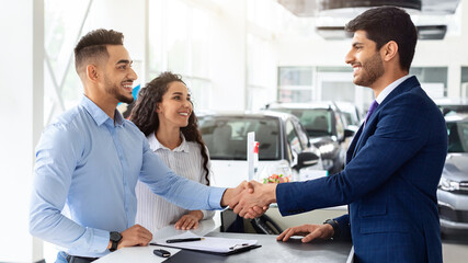 Smiling middle-eastern young man and woman buying car at auto salon, handsome arab guy shaking sales assistant hand, making successful deal, happy family got brand new luxury car, side view