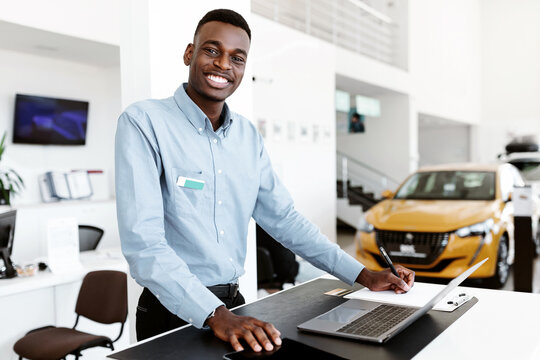 Portrait of happy car salesman standing at work desk, smiling at camera, using laptop and touch pad in auto dealership. Positive black automobile manager posing at workplace in showroom store