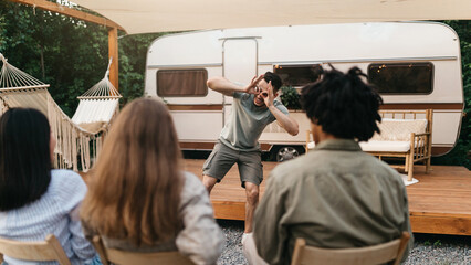 Multiracial young friends playing charades near motorhome during outdoor party. Millennial diverse...