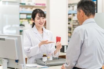 薬局のレジで薬の説明をする薬剤師の女性と高齢者男性
