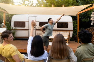 Diverse young friends playing charades near camper van, trying to guess word during outdoor party....