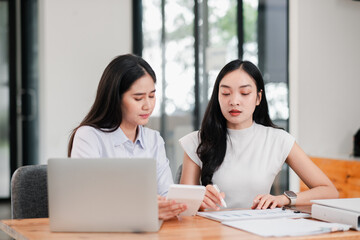 Obraz premium Two women working together in a contemporary office, discussing documents and using a laptop, showcasing teamwork and collaboration.