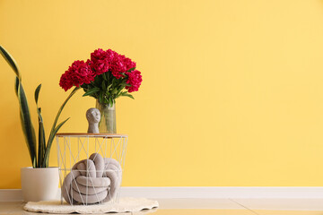 Vase of red peonies with figurine and houseplant near yellow wall