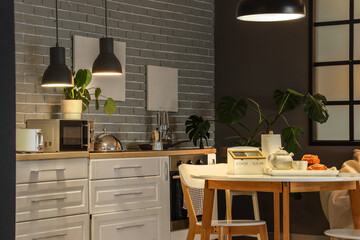 Interior of modern kitchen with glowing lamps, dining table and white counters at evening