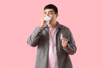 Ill young man with nasal drops and tissue on pink background