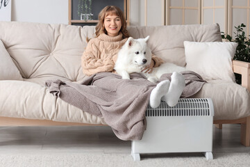 Young woman with blanket and Samoyed dog on sofa warming near radiator at home
