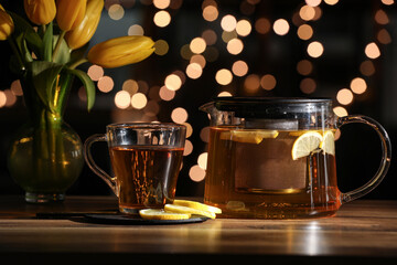 Teapot, glass of hot brewed tea and yellow tulips on wooden table against blurred background