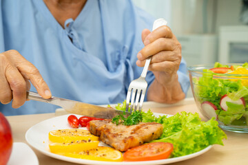 Asian senior woman patient eating pork chop stake and vegetable salad for healthy food in hospital.