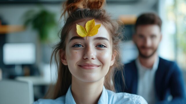 Cheerful young woman in office setting with yellow leaf on forehead, smiling playfully at camera while colleagues work in background. - Powered by Adobe