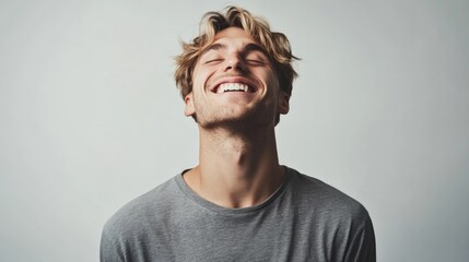 Obraz premium Carefree young man with blond wavy hair laughing joyfully in grey t-shirt against a soft white background smiling with closed eyes