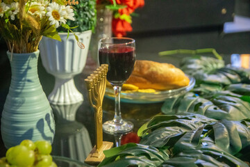 A wooden menorah stands on a table with a glass of red wine and a loaf of bread. Surrounding the menorah are green leaves and a white vase filled with flowers. The background features red flowers