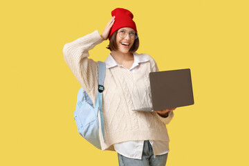 Beautiful young woman with modern laptop and backpack on yellow background