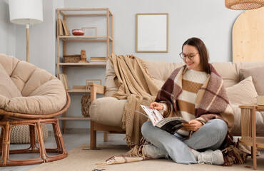 Fototapeta premium Young woman with warm plaid reading magazine on carpet in living room