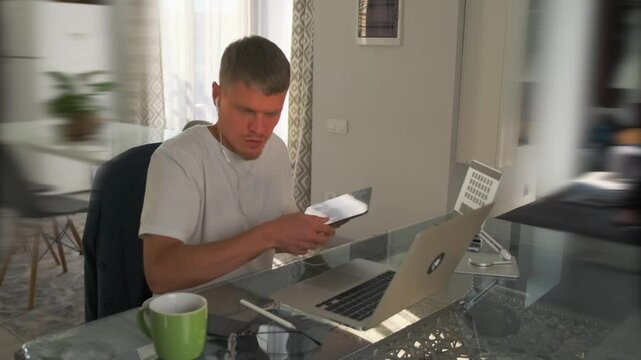 The developer, sitting at a desk with a laptop in the living room, picks up a tablet containing technical documentation and uses a stylus to highlight the necessary changes for improving the project.