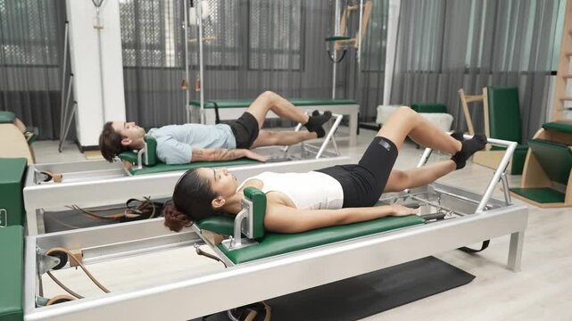 Couple practicing leg exercises on pilates reformer to strengthen core and improve flexibility at gym. Pilates partner doing pelvic bridge pose on reformer for improving lower back muscle. Habituate.