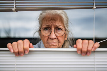 Senior woman stares out suspiciously through her blinds, her fingers separating them