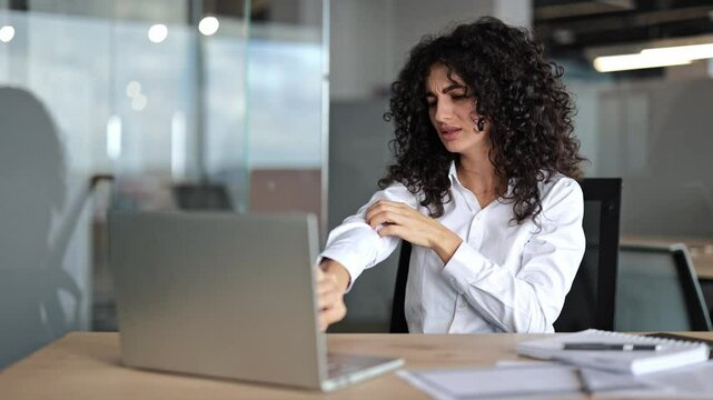 Stressed businesswoman with curly hair scratches her arm from apparent dry skin and discomfort in the office.
