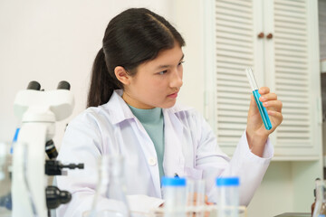 Fototapeta premium Cute Caucasian schoolgirl wearing white scientist outfit holding test tube filled with blue liquid in science classroom. Curious expression, analyzing chemical reaction, lab equipment and microscope.