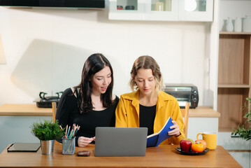 Two professional women collaborating on a business project with a laptop, notebook, and coffee at home.
