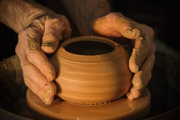 Potter's hands while working on the wheel, side view with dark background. Master makes a pot on a potter's wheel