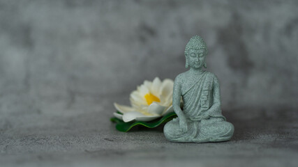 A statue of a Buddha sitting on a grey surface with a white flower in front of it. The statue is small and the flower is delicate. Concept of peace and tranquility