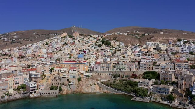 Discover an establishing panoramic aerial view of the Church of Agios Nikolaos at Asteria Beach, Syros Island, Greece. See the town sprawl across the landscape in a stunning Greek cityscape.