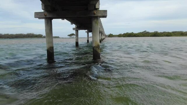 Rustic wooden bridge crossing tranquil Bellinger River, lagoon, Urunga, New South Wales, Australia. Aerial drone flying under and over footbridge