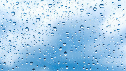 Water drops on glass with blue sky and white clouds as background.
