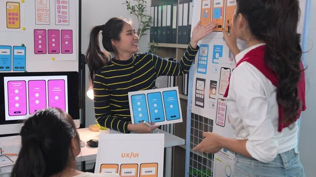 Female web designers collaborating on mobile app ui/ux design, analyzing layouts and functionalities using printed mockups and whiteboard in professional workspace