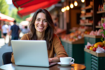 a woman, early 30s, sitting at a bistro table in an outdoor market with a laptop and cup of tea. digital nomad life.