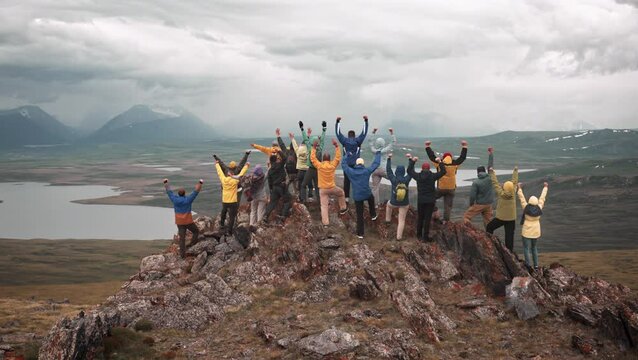 Big group of happy tourists are standing at mountain top with great view and celebrating finish of climbing to mountain top. Huge company of hikers are standing with open arms in winner pose