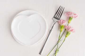 Top view of empty ceramic heart shaped plate served on white wooden background with pink carnation flowers and fork prepared for engagement proposal celebration or valentines day holiday greeting