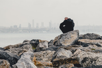 Contemplative man sits on rocks overlooking a hazy cityscape and calm water. A sense of solitude...