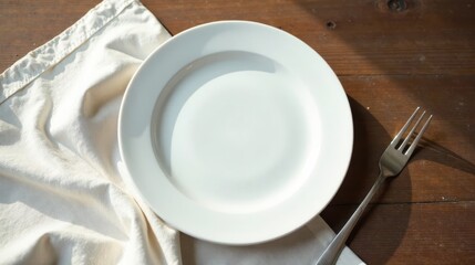 Empty white dinner plate on a linen cloth with a fork on a wooden table bathed in sunlight